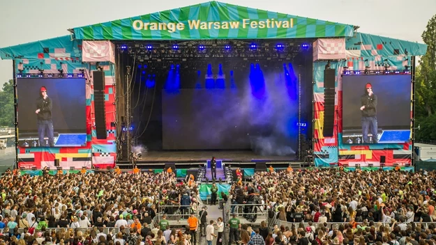 Duża scena festiwalowa Orange Warsaw Festival, tłum ludzi zgromadzonych przed sceną, na scenie wykonawca przemawia do publiczności, po bokach ekrany wyświetlające obraz sceny, nad całością dominują kolorowe elementy scenografii.