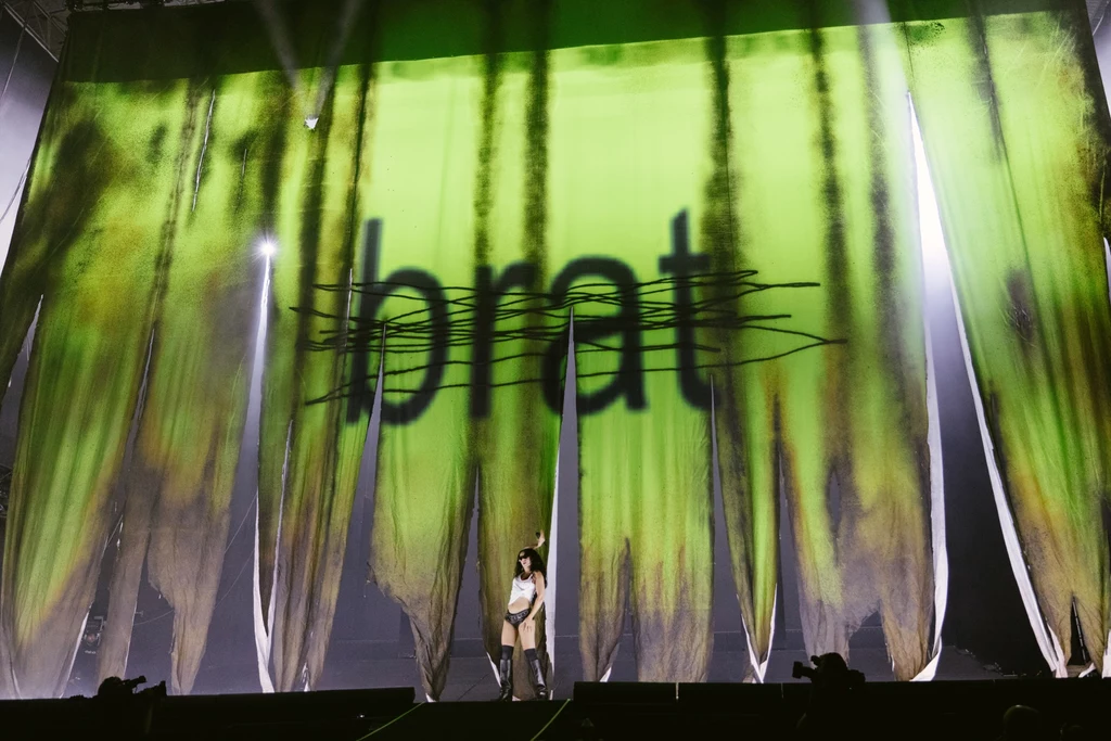 Charli xcx na Orange Warsaw Festival Osoba stojąca na scenie z dużymi zielonymi kotarami, na których widnieje czarne, przekreślone słowo 'brat', tło podświetlone jest efektami świetlnymi.